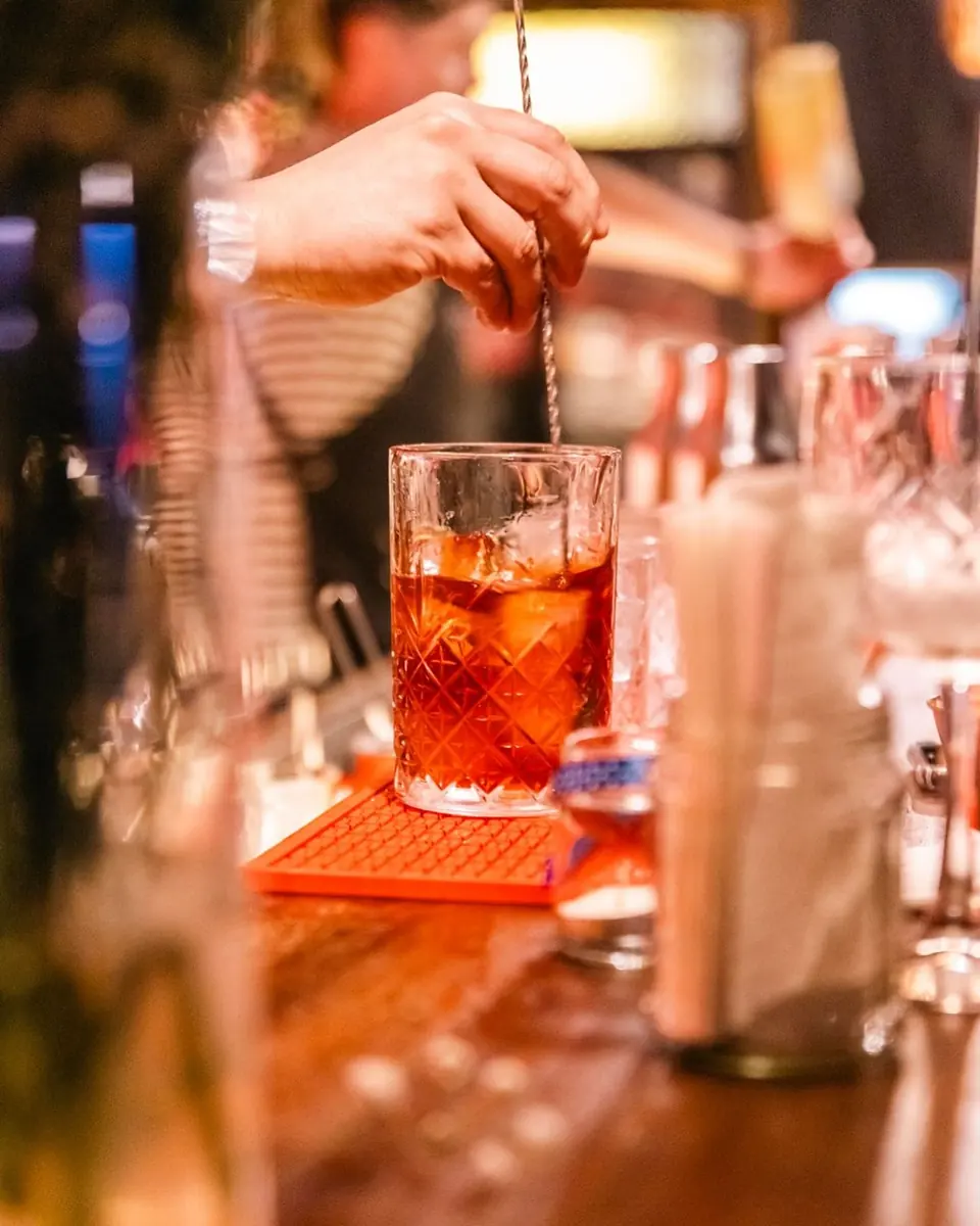 Un bartender revuelve un trago rojizo con hielo en la barra, luz ámbar