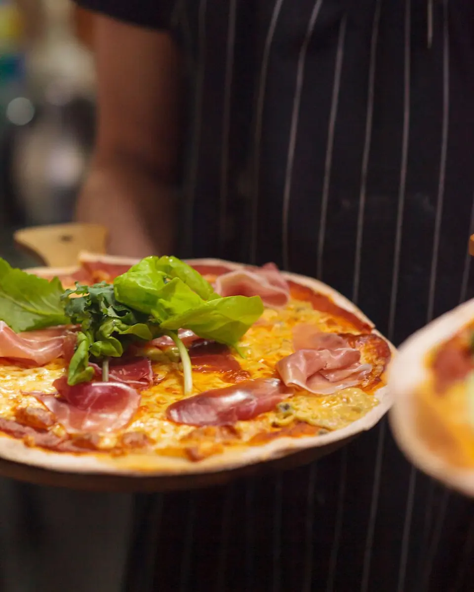 Un garzón lleva una pizza de masa delgada con jamón serrano y rúcula