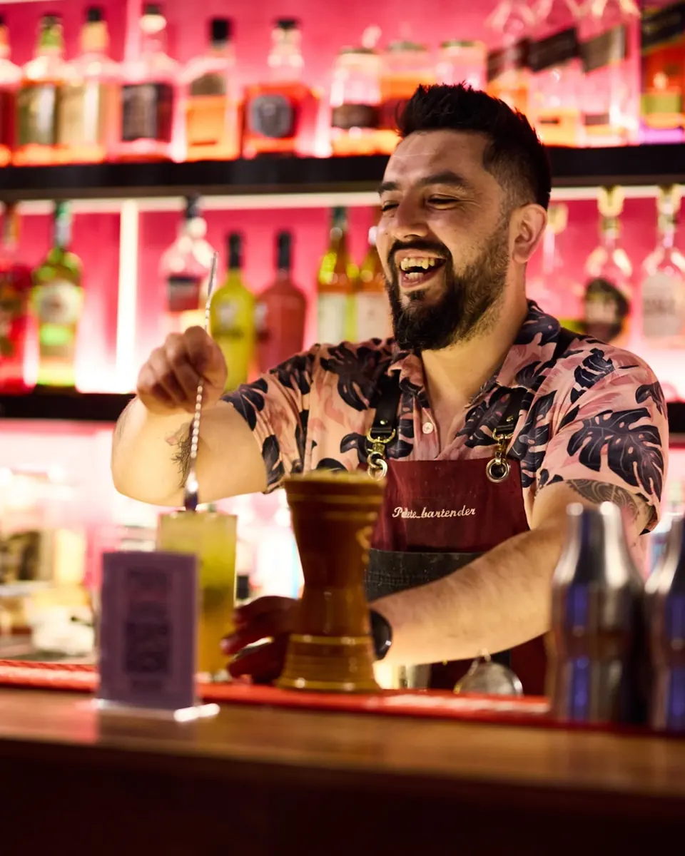 Bartender riendo con camisa floreada frente al muro de botellas rosado