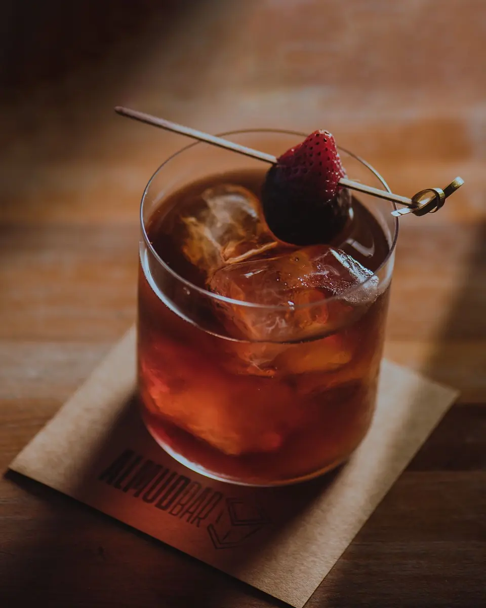 Trago rojizo con hielo y una frutilla sobre un posavasos de Almud