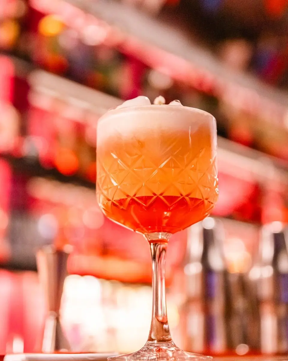 Copa tallada con un trago naranja y espuma, con las botellas rosadas desenfocadas atrás